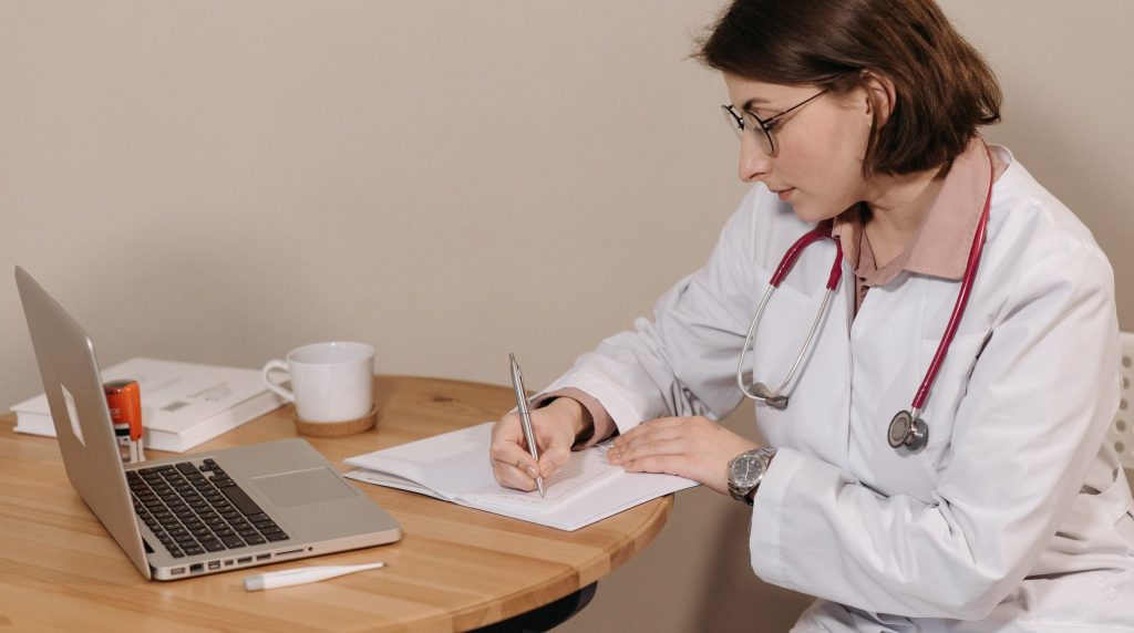 médecin en blouse rédigeant un document à la main à son bureau, ordinateur portable à côté